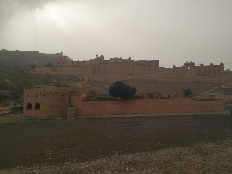 A sandstone fortress complex rising over dusty hills under hazy sun